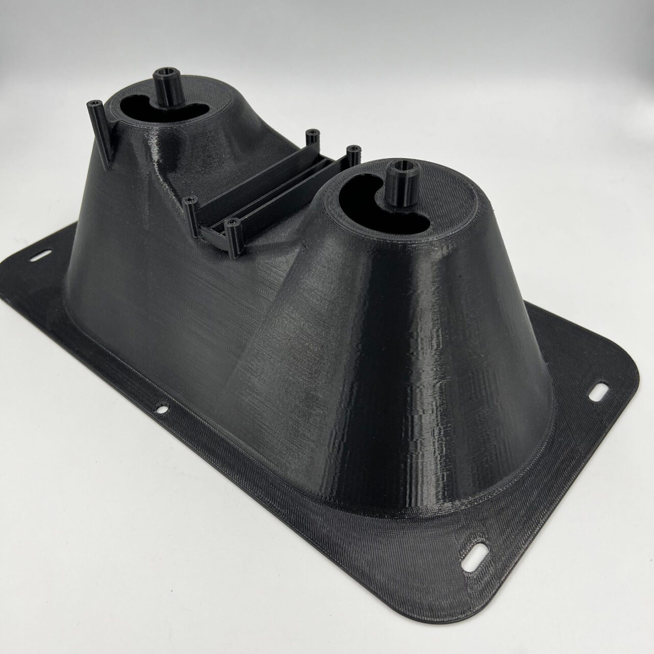 A black, 3D-printed plastic component crafted using fused deposition modeling features two cylindrical protrusions and a central rectangular section. The base is rectangular with rounded corners, complete with small mounting holes along the edges.