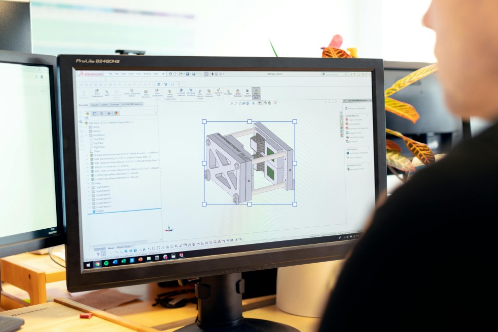 A person uses CAD software on a computer to design a 3D mechanical component for additive manufacturing. The monitor displays a detailed model of a metal frame with various parts and technical drawings.