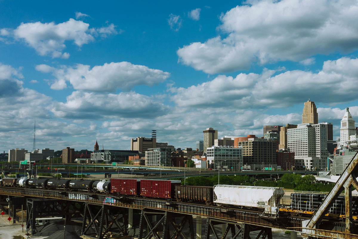 A skyline of a city with tall buildings under a blue sky and scattered clouds. In the foreground, an Ohio Manufacturing freight train travels on elevated tracks, carrying tankers and boxcars above highways and greenery.