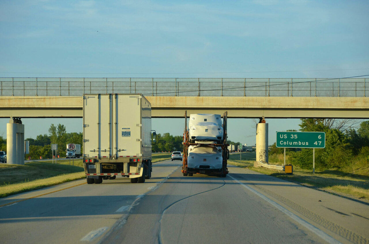 A highway scene with two trucks driving under an overpass; one truck transports semi-truck cabs—showcasing Ohio manufacturing. A green road sign marks US 35, Route 6, and Columbus, 47 miles away. Trees line the road on a clear day.