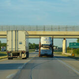 A highway scene with two trucks driving under an overpass; one truck transports semi-truck cabs—showcasing Ohio manufacturing. A green road sign marks US 35, Route 6, and Columbus, 47 miles away. Trees line the road on a clear day.